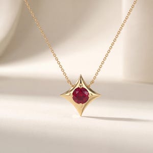 May include: A gold necklace featuring a four-pointed star-shaped pendant with a round, deep red gemstone set in the center. The pendant is suspended from a delicate gold chain, showcasing a polished metallic finish and a symmetrical design.