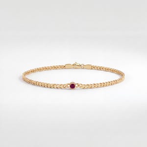 May include: A gold chain bracelet featuring a single round, bezel-set red gemstone centered along the curb link chain. The bracelet is finished with a secure lobster claw clasp mechanism, showcasing a polished metallic texture and vibrant color.