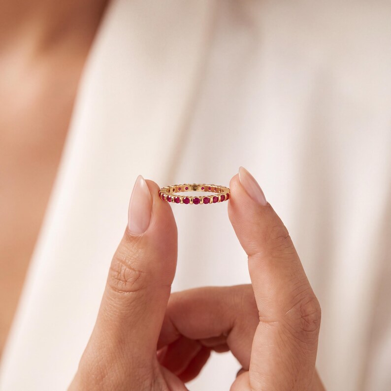 K&ouml;nnte beinhalten: Ein goldener Eternity-Ring mit einer durchgehenden Reihe kleiner, runder, leuchtend roter Rubine, die in einen polierten Metallrahmen gefasst sind. Der Ring ist schmal und zierlich und zeigt ein gleichm&auml;&szlig;iges Design mit sicher gefassten Steinen.