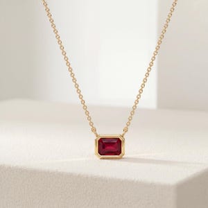 May include: A gold chain necklace featuring a single emerald-cut red gemstone pendant set in a polished gold bezel. The pendant is centered on a delicate gold cable chain, showcasing a vibrant red stone with clean, geometric lines and a warm metal finish.