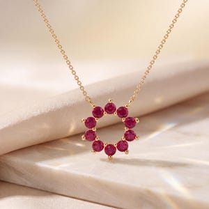 Puede incluir: Un collar de cadena dorada con un colgante circular compuesto por diez piedras preciosas redondas de color rojo vibrante dispuestas en forma de corona. El engaste es de oro pulido y las piedras están sujetas por pequeñas garras finas.
