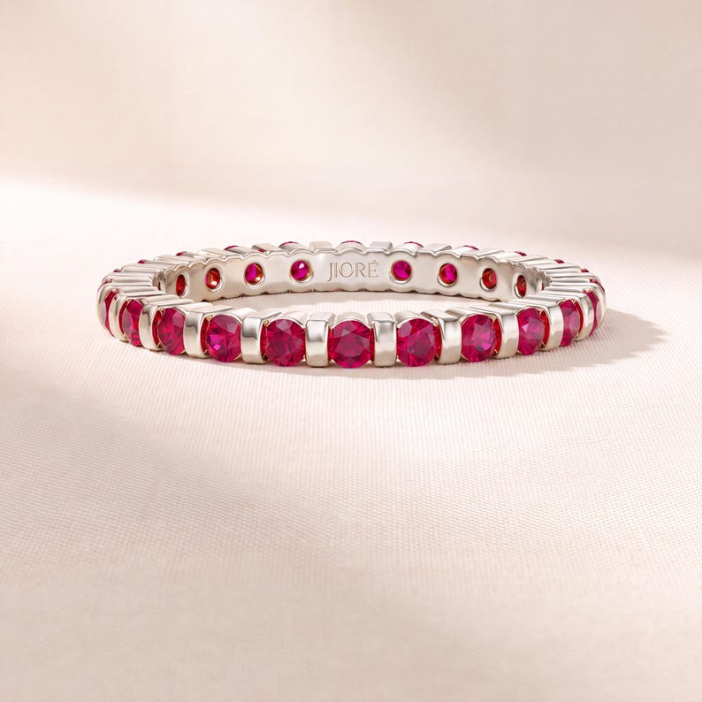 K&ouml;nnte beinhalten: Ein silberfarbener Eternity-Ring mit einer durchgehenden Reihe runder, leuchtend roter Steine in einer Stegfassung. Die Innenseite des Rings ist mit dem Markennamen JIOR&Eacute; in einer klaren, serifenlosen Schrift graviert, die am Innenrand sichtbar ist.