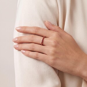K&ouml;nnte beinhalten: Ein d&uuml;nner, zierlicher Ring mit einem durchgehenden Band aus kleinen, runden, tiefroten Edelsteinen in einer Metallfassung. Der Ring wird am Ringfinger getragen und zeigt ein schlichtes Design mit einer gleichm&auml;&szlig;igen Anordnung der Steine.