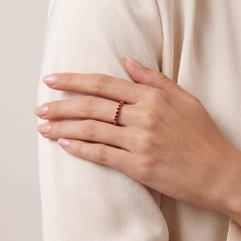 K&ouml;nnte beinhalten: Ein d&uuml;nner, zierlicher Ring mit einem durchgehenden Band aus kleinen, runden, tiefroten Edelsteinen in einer Metallfassung. Der Ring wird am Ringfinger getragen und zeigt ein schlichtes Design mit einer gleichm&auml;&szlig;igen Anordnung der Steine.