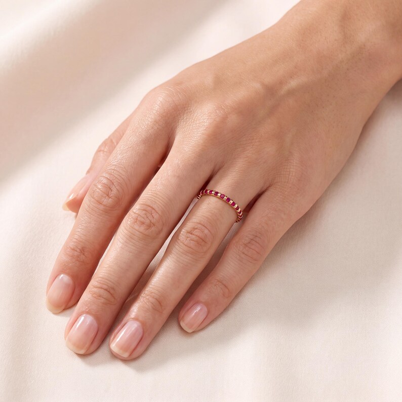 K&ouml;nnte beinhalten: Ein d&uuml;nner Goldring mit einer durchgehenden Reihe kleiner, runder, leuchtend roter Edelsteine, die rund um den gesamten Ring gefasst sind. Der Ring wird am Ringfinger getragen und zeigt die polierte Metalloberfl&auml;che sowie die gleichm&auml;&szlig;igen Steine.