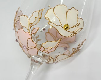 Puede incluir: Una copa de vino de vidrio transparente con un diseño floral rosa y blanco pintado en el lateral. El diseño presenta detalles dorados y pequeños diamantes de imitación morados. La copa tiene el texto "BOHEMIA SELECTION" en la parte superior.