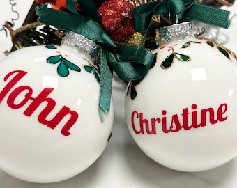 May include: Two white Christmas ornaments with green holly leaf designs. One ornament says "John" in red lettering and the other says "Christine" in red lettering.