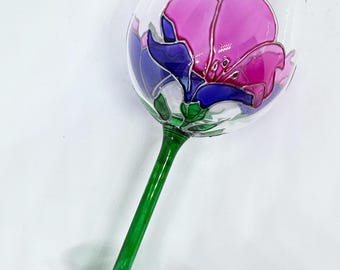 Puede incluir: Una copa de vino pintada a mano con un diseño floral. La copa presenta una flor rosa y morada con detalles azules, delineada en plata, sobre una copa transparente. Tiene un tallo y base verdes.