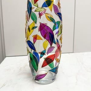 Peut inclure: Un vase en verre transparent avec un motif de feuilles colorées. Les feuilles sont peintes dans des tons de rouge, jaune, bleu, vert et violet, soulignées d'or. Le vase est cylindrique et repose sur une surface blanche.