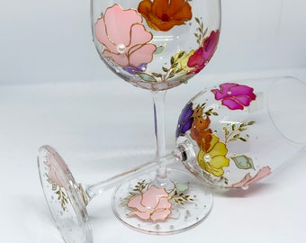 Puede incluir: Dos copas de vino de vidrio transparente con diseños florales pintados a mano. Las copas presentan flores rosas, naranjas, amarillas, moradas y verdes con detalles dorados y adornos de perlas blancas.