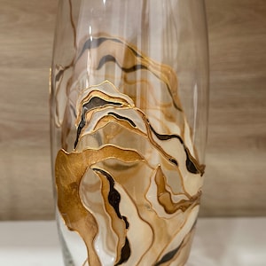Può includere: Un vaso di vetro trasparente con un design astratto dorato e nero. Il design presenta linee e forme vorticose.