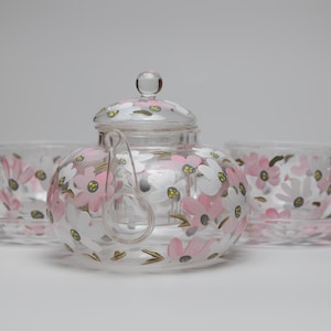 Könnte beinhalten: Ein klares Glas-Teeservice mit einer Teekanne, zwei Tassen und Untertassen. Das Set ist mit rosa und weißen Blumenmustern verziert. Die Teekanne hat einen Deckel und einen klaren Glas-Teefilter.