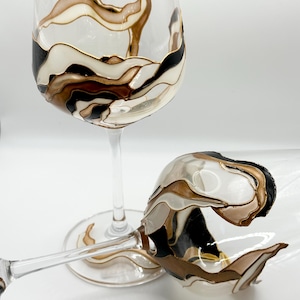 Puede incluir: Dos copas de vino de vidrio transparente con diseños pintados abstractos. Las copas presentan remolinos negros, blancos y dorados.