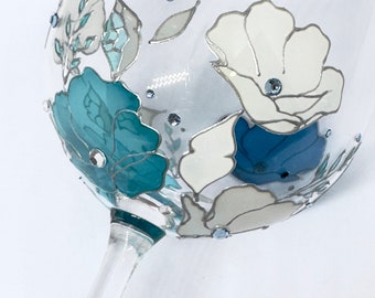Puede incluir: Una copa de vino de vidrio transparente con un diseño pintado de flores blancas y azules. Las flores están delineadas en negro y tienen pequeños diamantes de imitación azules esparcidos por todo el diseño.