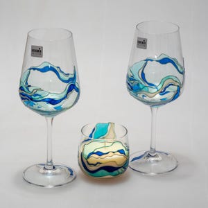 Puede incluir: Dos copas de vino y un pequeño portavelas. Cada pieza es de vidrio transparente con un diseño de olas azules, turquesas y blancas. Las copas de vino tienen tallos largos y el portavelas es un vaso pequeño y redondo. La etiqueta de las copas dice "Bohemia Selection."