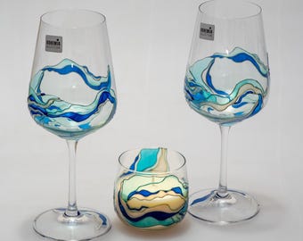 Puede incluir: Dos copas de vino y un pequeño portavelas. Cada pieza es de vidrio transparente con un diseño de olas azules, turquesas y blancas. Las copas de vino tienen tallos largos y el portavelas es un vaso pequeño y redondo. La etiqueta de las copas dice "Bohemia Selection."