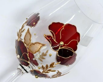 Puede incluir: Una copa de vino de vidrio transparente con un diseño pintado a mano de amapolas rojas y hojas doradas. La copa tiene un tallo delicado y una copa redonda.