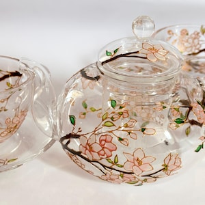Pode incluir: Um bule de vidro transparente e duas chávenas a condizer com pires, decorados com flores de cerejeira cor-de-rosa pintadas à mão sobre ramos castanhos com pequenas folhas verdes. O bule inclui uma tampa de vidro com um puxador redondo.