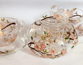 May include: A clear glass teapot and two matching teacups with saucers, each decorated with hand-painted pink cherry blossom flowers on brown branches with small green leaves. The teapot features a glass lid with a round knob handle.