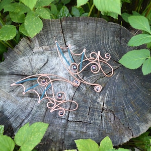 May include: Two copper-colored wire hair accessories with an ornate, swirling design. Each piece features a series of small, iridescent beads and teal-colored accents. The accessories are displayed on a weathered, gray tree stump, surrounded by green foliage.