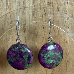 Könnte beinhalten: Ein Paar Ohrhänger mit runden, polierten Steinen in tiefen Lila- und Grüntönen. Die Ohrringe haben silberfarbene Klappbügelverschlüsse.