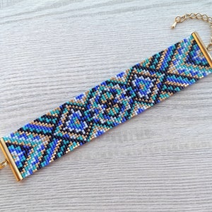 May include: A wide beaded bracelet with a geometric pattern in shades of blue, teal, black, and gold. The bracelet has gold-colored metal end caps and a matching chain clasp for adjustable sizing. The bracelet is approximately 1.5 inches wide.