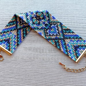 May include: A beaded bracelet with a geometric pattern in shades of blue, teal, black, and gold. The bracelet has gold-colored metal end caps and a matching chain with a lobster clasp for adjustable sizing. The bracelet is approximately 1.5 inches wide.