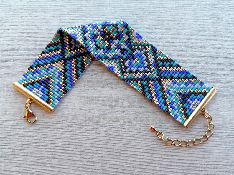 May include: A beaded bracelet with a geometric pattern in shades of blue, teal, black, and gold. The bracelet has gold-colored metal end caps and a matching chain with a lobster clasp for adjustable sizing. The bracelet is approximately 1.5 inches wide.