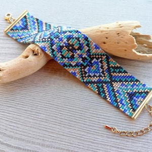 May include: A wide beaded bracelet with a geometric pattern in shades of blue, black, white, and gold. The bracelet has gold-colored metal end caps and a chain clasp. The bracelet is resting on a piece of driftwood.