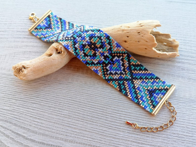 May include: A wide beaded bracelet with a geometric pattern in shades of blue, black, white, and gold. The bracelet has gold-colored metal end caps and a chain clasp. The bracelet is resting on a piece of driftwood.