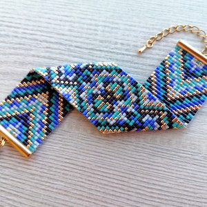 May include: A beaded bracelet with a geometric pattern in shades of blue, teal, black, and white. The bracelet has gold-colored metal end caps and a matching chain for adjustable sizing. The clasp is also gold-colored.