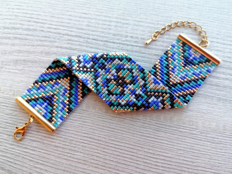 May include: A beaded bracelet with a geometric pattern in shades of blue, teal, black, and white. The bracelet has gold-colored metal end caps and a matching chain for adjustable sizing. The clasp is also gold-colored.