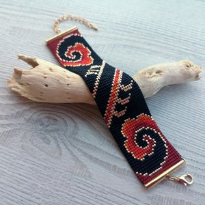 K&ouml;nnte beinhalten: Ein schwarzes und rotes Perlenarmband mit einem goldenen Verschluss. Das Armband hat ein geometrisches Muster mit Spiralen und Linien.