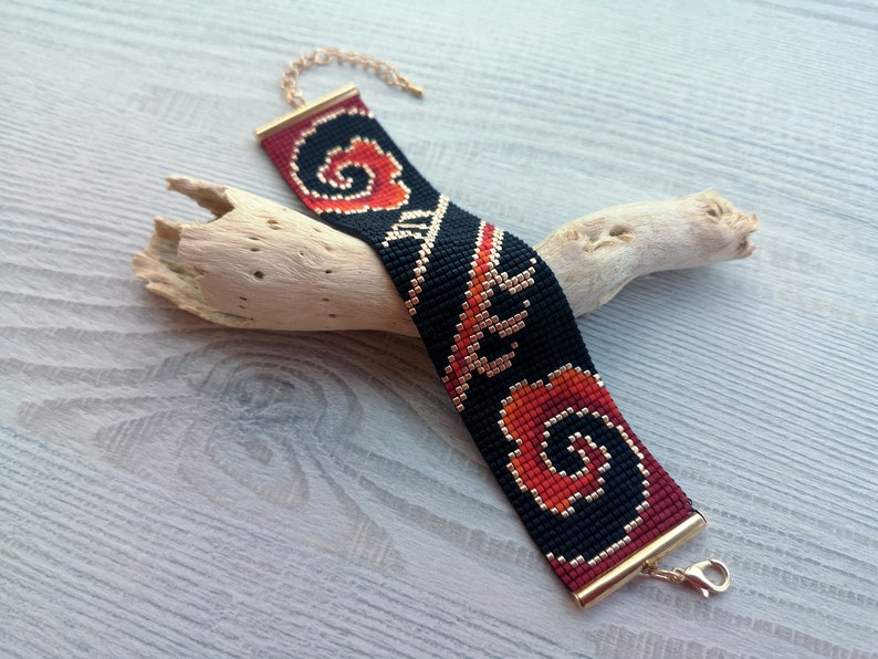 K&ouml;nnte beinhalten: Ein schwarzes und rotes Perlenarmband mit einem goldenen Verschluss. Das Armband hat ein geometrisches Muster mit Spiralen und Linien.