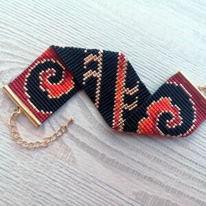 K&ouml;nnte beinhalten: Ein schwarzes, rotes und goldenes Perlenarmband mit einem Kettenverschluss. Das Armband hat ein geometrisches Muster.