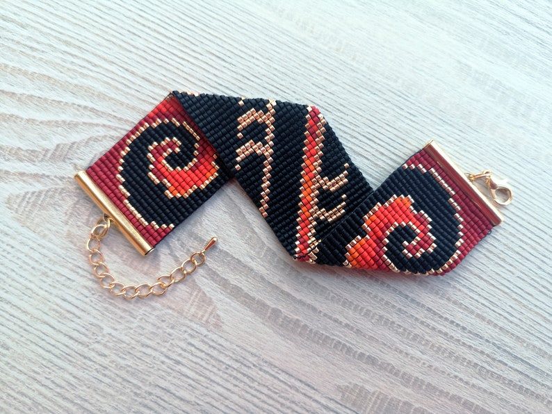 K&ouml;nnte beinhalten: Ein schwarzes, rotes und goldenes Perlenarmband mit einem Kettenverschluss. Das Armband hat ein geometrisches Muster.