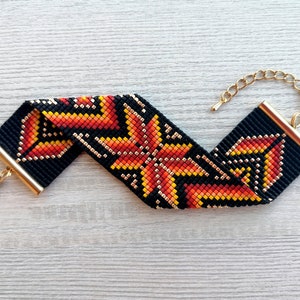 May include: A black beaded bracelet with a geometric pattern in orange, yellow, and gold. The bracelet has a gold clasp.
