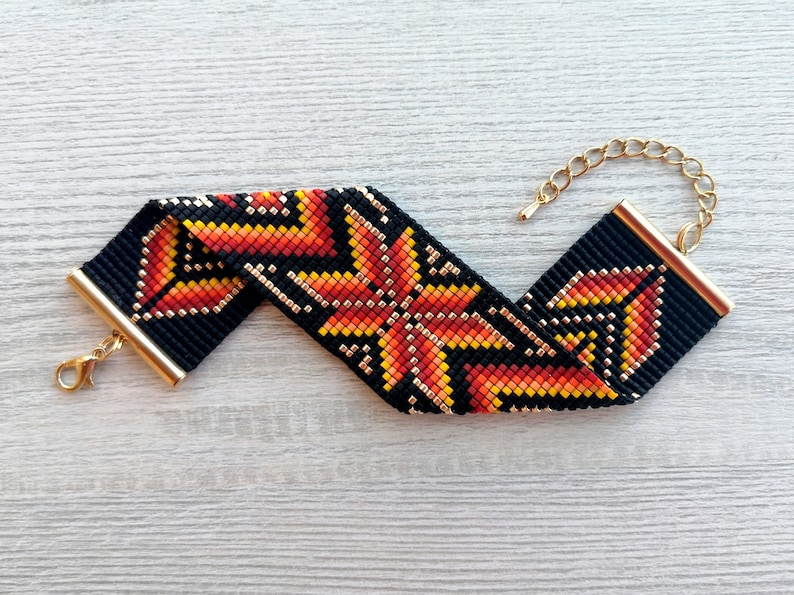 May include: A black beaded bracelet with a geometric pattern in orange, yellow, and gold. The bracelet has a gold clasp.