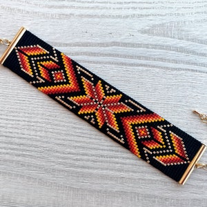 May include: A black beaded bracelet with a geometric pattern in shades of orange, yellow, and gold. The bracelet has a gold clasp and extender chain.