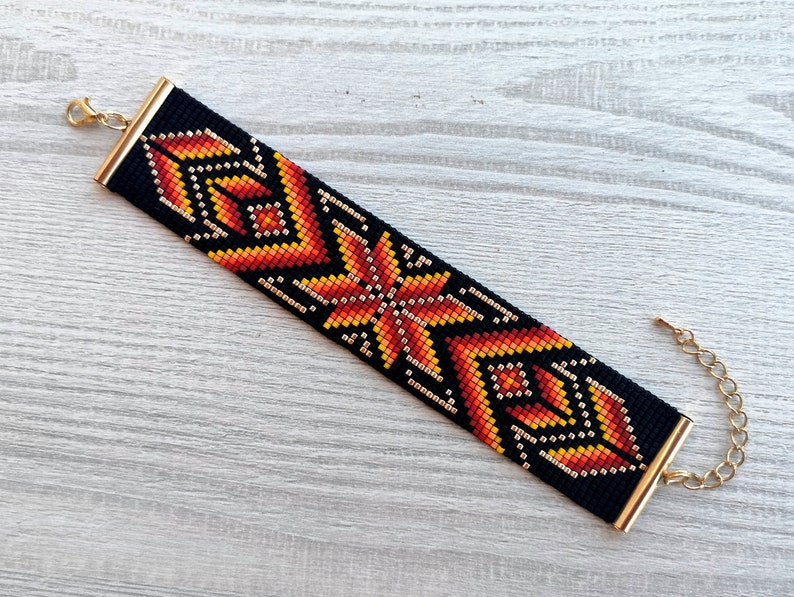May include: A black beaded bracelet with a geometric pattern in shades of orange, yellow, and gold. The bracelet has a gold clasp and extender chain.