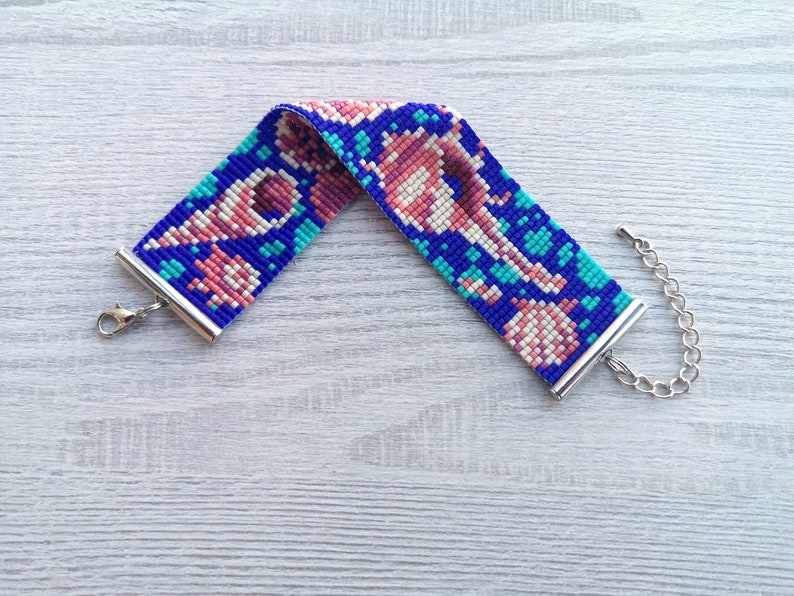 May include: A beaded bracelet with a colorful geometric pattern in shades of pink, blue, and white. The bracelet has silver metal clasps.