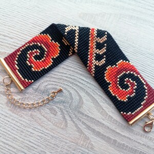 K&ouml;nnte beinhalten: Ein schwarzes und rotes Perlenarmband mit einem goldenen Verschluss und einer Kette. Das Armband hat ein geometrisches Muster mit Wirbeln.