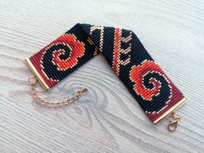 K&ouml;nnte beinhalten: Ein schwarzes und rotes Perlenarmband mit einem goldenen Verschluss und einer Kette. Das Armband hat ein geometrisches Muster mit Wirbeln.