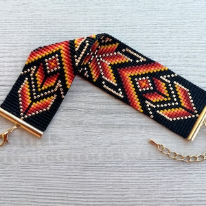 May include: A black beaded bracelet with a geometric pattern in orange, red, and gold. The bracelet has a gold clasp and chain.