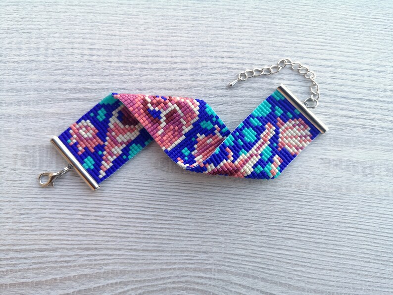 May include: A beaded bracelet with a blue, pink, and white geometric pattern. The bracelet has a silver clasp and chain.