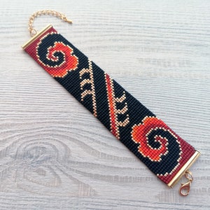 K&ouml;nnte beinhalten: Ein schwarzes Perlenarmband mit einem roten und orangefarbenen Blumenmuster und goldenen Akzenten. Das Armband hat einen goldenen Verschluss.