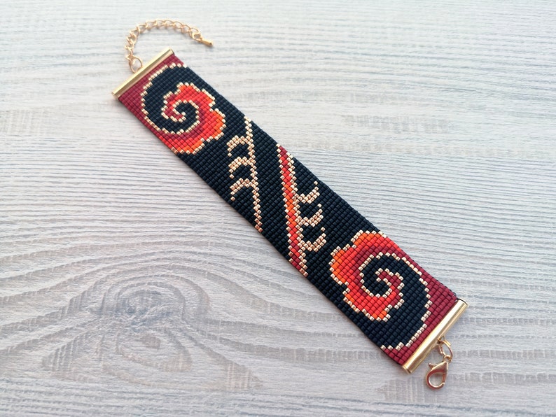 K&ouml;nnte beinhalten: Ein schwarzes Perlenarmband mit einem roten und orangefarbenen Blumenmuster und goldenen Akzenten. Das Armband hat einen goldenen Verschluss.