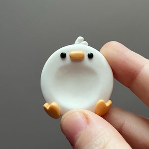 Op de afbeelding: Een wit keramiek kuikenbeeldje met zwarte ogen en een gele snavel. Het kuiken heeft oranje pootjes en zit op een hand.
