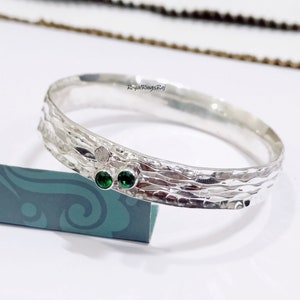 Puede incluir: Pulsera de plata con textura martillada y dos piedras preciosas verdes. La pulsera está sobre una superficie con estampado azul y verde. "RoyalRingsRaj" está impreso en la pulsera.
