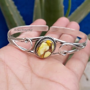 May include: A silver cuff bracelet featuring a central oval-shaped yellow and brown stone set in a twisted metal border. The band has open ends and decorative swirling metal wire accents on both sides. The text RoyalRingRaj is visible on the metal.